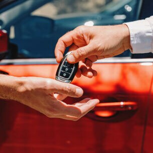 Eine Hand übergibt einen Autoschlüssel in eine andere Hand. Im Hintergrund ist ein rotes Auto zu sehen