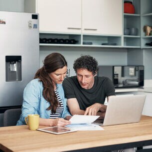 Ein junges Paar sitzt in der Küche mit einem Laptop, einem Tablet und einigem Papierkram.