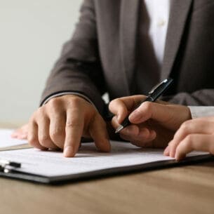Zwei Personen in Businesskleidung über einem Dokument; die eine Person deutet auf eine Passage, während die andere einen Kugelschreiber hält.