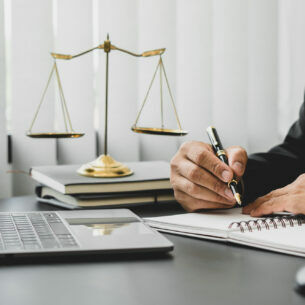 Person im Anzug mit Laptop, Block und Stift bei der Arbeit am Schreibtisch, im Hintergrund steht die Waage der Gerechtigkeit