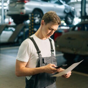 Eine Person in Arbeitskleidung steht in einer Werkstatt und hält ein Smartphone sowie ein Dokument in ihren Händen
