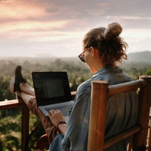 Eine Frau sitzt im Freien auf einer Terrasse, arbeitet an einem Laptop mit Blick ins Grüne.