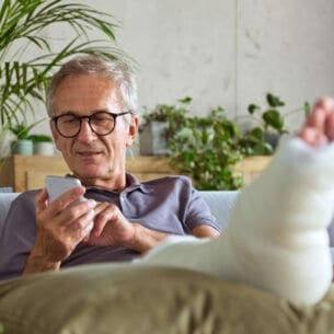 Ein Mann auf einem Sofa, der ein Smartphone in der Hand hält und seinen eingegipsten Fuß hochlegt