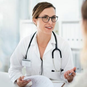 Eine weibliche Person mit Brille im Arztkittel spricht mit einer anderen Person