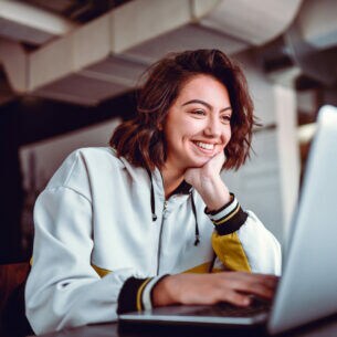 Eine junge Studentin sitzt lächelnd vor einem Laptop in einem Universitätsgebäude.