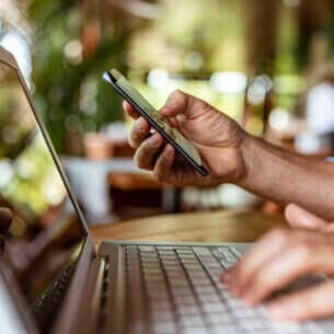 Arm einer Person, die vor einem Laptop sitzt und ein Smartphone in der rechten Hand hält, während er mit der linken Hand die Laptop-Tastatur bedient.