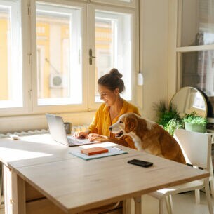 Eine Frau sitzt an einem Tisch und arbeitet am Laptop, neben ihr sitzt ein Hund
