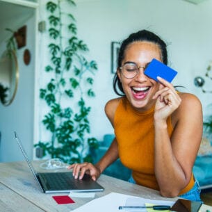 Eine junge Frau mit Brille sitzt lachend vor einem aufgeklappten Laptop und hält sich eine Bankkarte vors Gesicht.