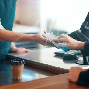 Eine Person, die über einen Tresen, auf dem ein Kaffeebecher steht, Zahlen in ein Terminal eingibt, das eine Person auf der anderen Seite des Tresens hinhält