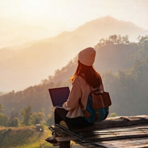 Eine Person mit Laptop sitzt auf einem hölzernen Untergrund und blickt auf ein Naturpanorama mit Bergen und Sonnenaufgang