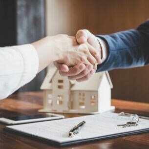 Zwei sich schüttelnde Hände über einem Dokument, im Hintergrund steht ein Modellhaus aus Holz