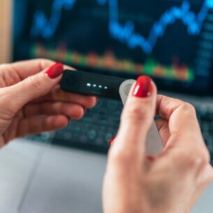 Weibliche Hände halten eine Hardware Wallet vor einem Laptop hoch
