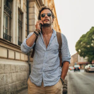 Mann mit Bart, Sonnenbrille und Rucksack auf dem Rücken steht auf einem Bürgersteig und telefoniert