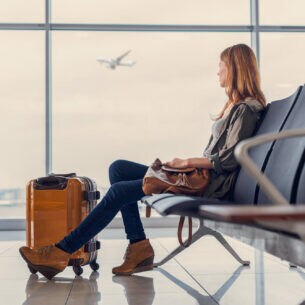 Eine Person sitzt mit einem Koffer im Flughafen und blickt aus dem Fenster, an dem gerade ein Flugzeug vorbeifliegt
