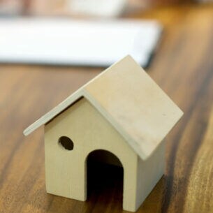 Ein kleines Hausmodell aus Holz steht auf einem Schreibtisch, im Hintergrund sind unscharf zwei Personen und Unterlagen zu erkennen.