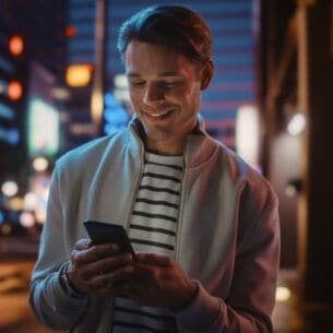 Ein Mann steht auf der Straße einer nächtlich beleuchteten Stadt und schaut lächelnd auf das Smartphone in seiner Hand.