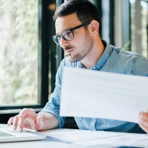 Eine Person sitzt an einem Tisch, hält ein Dokument in der einen Hand und tippt mit der anderen auf einem Notebook.