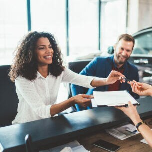 Zwei Personen sitzen im Autohaus am Schreibtisch einer anderen Person gegenüber und überreichen sich gegenseitig Dokumente und Autoschlüssel