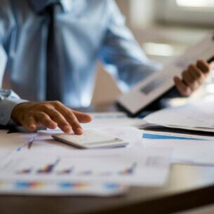 Eine Person sitzt am Schreibtisch und hat vor sich diverse Dokumente ausgebreitet und tippt auf einem Taschenrechner.