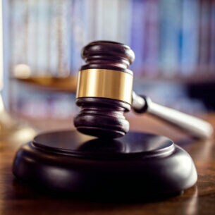 Auf einem Tisch liegt ein Richterhammer auf einem Resonanzblock. Im Hintergrund steht eine Waage.