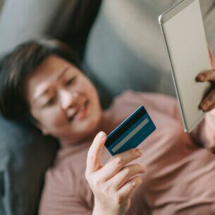 Eine Person liegt auf der Couch. Sie hält in der einen Hand eine Kreditkarte und in der anderen ein Smartphone.