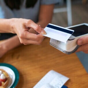 Eine Person hält eine Bankkarte an ein elektrisches Lesegerät, das eine andere Person hält. Auf einem Tisch darunter liegt ein Kassenbon.