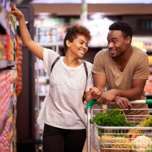 Zwei Personen zwischen Supermarktregalen. Sie schieben einen mit Gemüse gefüllten Einkaufswagen.