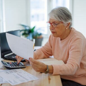 Eine ältere Dame an einem Tisch hält ein Blatt Papier in der Hand und tippt auf einen Taschenrechner.