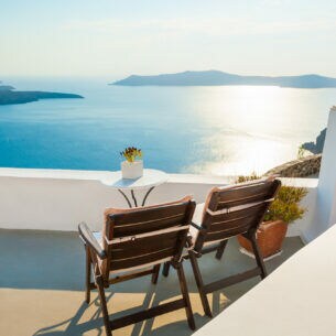 Zwei Holzstühle stehen auf einer sonnigen Terrasse mit Blick übers Meer.
