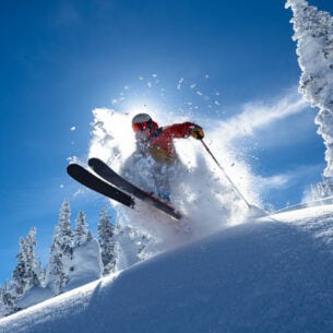 Eine Person springt mit seinen Skiern über einen kleinen Schneehügel, hinter ihm scheint die Sonne.