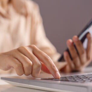 Eine Frau hält ein Handy in der linken Hand und drückt mit der rechten Hand eine Taste auf ihrem Laptop