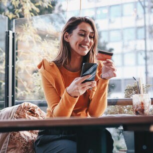 Eine Person sitzt an einem Tisch und hält lächelnd eine Kreditkarte in der Hand.