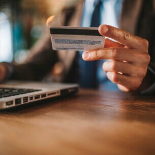 Nahaufnahme einer Hand, die neben einem geöffneten Laptop eine Kreditkarte in der Hand hält.