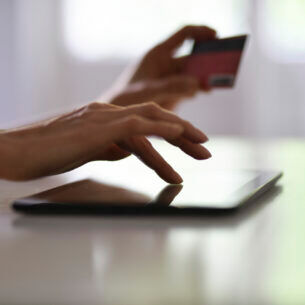 Nahaufnahme einer Hand, die auf einem Tablet tippt. Im Hintergrund ist eine zweite Hand zu erkenne, die eine Bankkarte in der Hand hält.