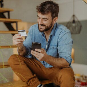 Ein Mann sitzt lächelnd auf den Stufen einer Treppe. In der einen Hand hält er eine Kreditkarte, auf die er schaut. In der anderen Hand hält er ein Smartphone