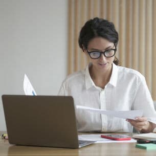 Eine Person sitzt am Schreibtisch mit einem Laptop und hält Zettel in der Hand