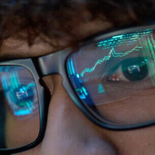 Eine Person schaut auf einen Monitor. In ihrer Brille spiegeln sich die Charts von Aktienkursen.