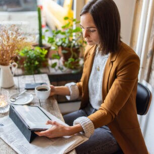 Eine Frau trinkt einen Kaffee und scrollt dabei durch Feed von Nachrichten auf ihrem Tablet.
