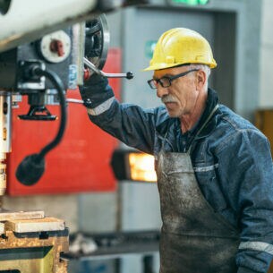 Ein älterer Mann mit gelbem Helm und Blaumann steht an einer Industriebohrmaschine.