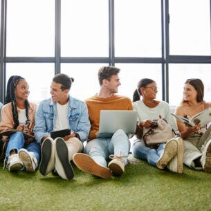 Mehrere junge Personen sitzen nebeneinander auf dem Fußboden an eine Fensterfront gelehnt.