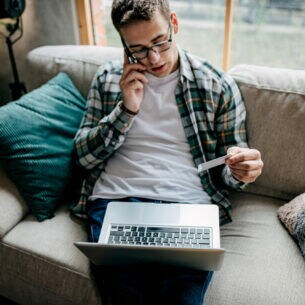 Eine Person aus der Aufsicht, die telefonierend auf einem Sofa sitzt, eine Bankkarte in der Hand hält und ein Laptop auf dem Schoß liegen hat.