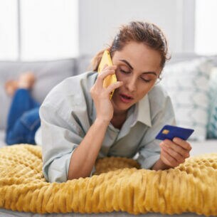 Eine Person liegt bäuchlings und telefonierend auf dem Sofa und hält eine Kreditkarte in der Hand.