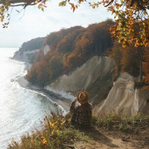 Eine Person mit großem Hut sitzt an einer Klippe mit Blick Richtung Meer.