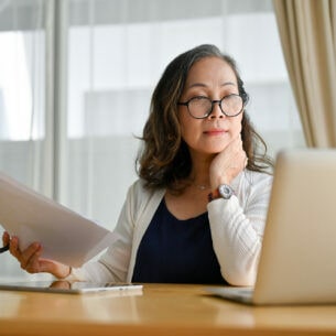 Eine Person mit Brille sitzt am Schreibtisch, blickt auf ihr Laptop und hält dabei Dokumente in der Hand.