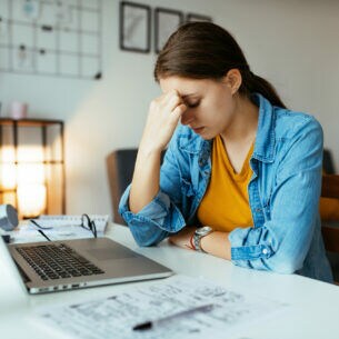 Eine Frau sitzt am Tisch vor einem Laptop und stützt den Kopf mit einer Hand.