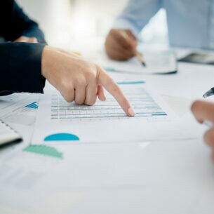Drei Personen an einem Tisch. Eine Person zeigt auf ein Papier in der Mitte, auf dem Tabellen abgebildet sind