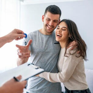 Zwei Personen umarmen sich und bekommen dabei von einer dritten Person einen Schlüssel überreicht.