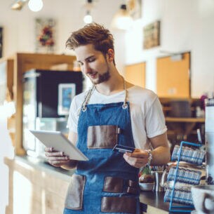 Ein junger Barista steht in einem Café, er hat eine Schürze an und hält in der einen Hand ein Tablet und in der anderen Hand eine Kreditkarte