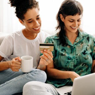 Zwei auf dem Sofa sitzende Personen halten eine Tasse und eine Kreditkarte in der Hand. Eine Person hat einen Laptop auf dem Schoß.