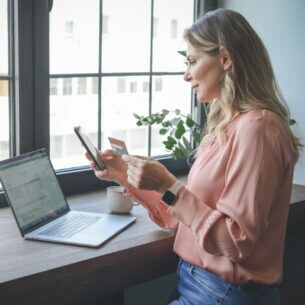 Eine Frau steht mit Handy und Kreditkarte in der Hand vor einem Tisch, auf dem ein Laptop steht